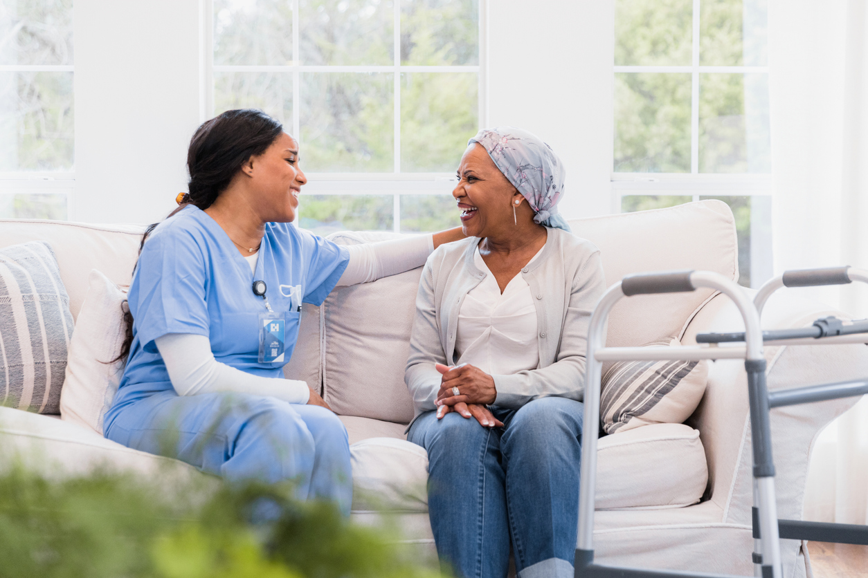 A smiling nurse and a hospice patient sit together on a couch.
