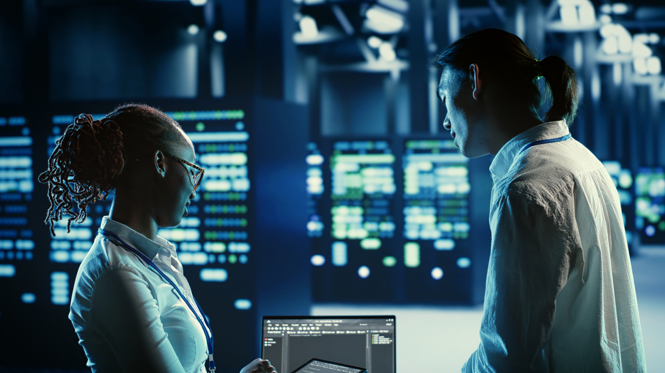 Two cybersecurity professionals standing in a server room read a laptop screen.