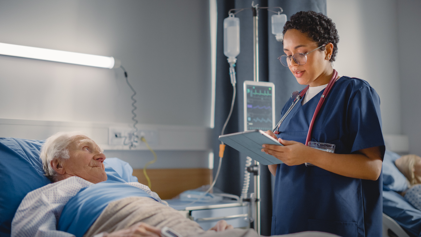 A clinical nurse specialist holding a tablet assesses a patient in a hospital bed.