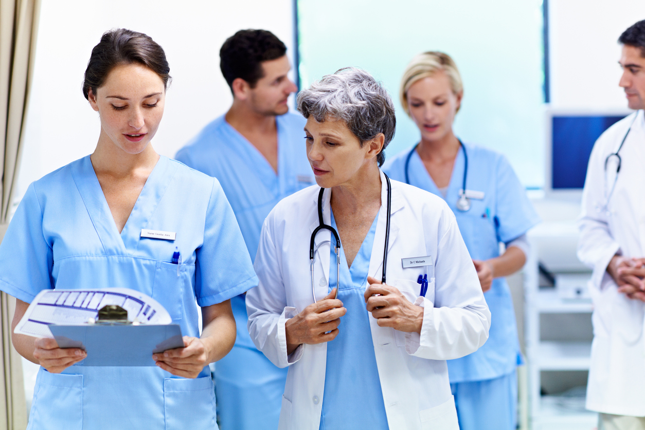 A physician talks with a charge nurse holding a clipboard in a busy hospital room.