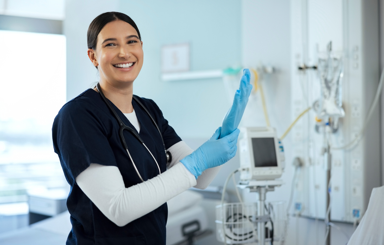 A smiling nurse puts on a latex glove in a hospital room.
