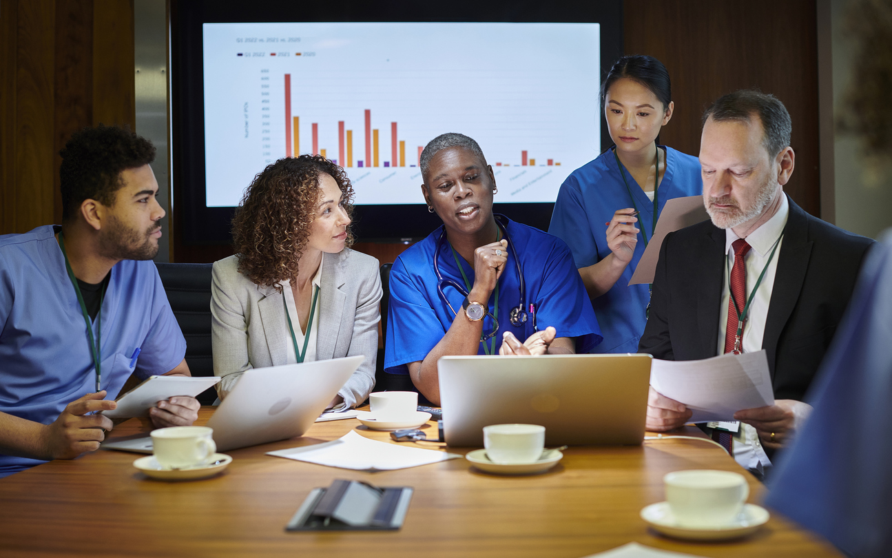 Nurses and administrators meet around a conference table in front of a monitor.