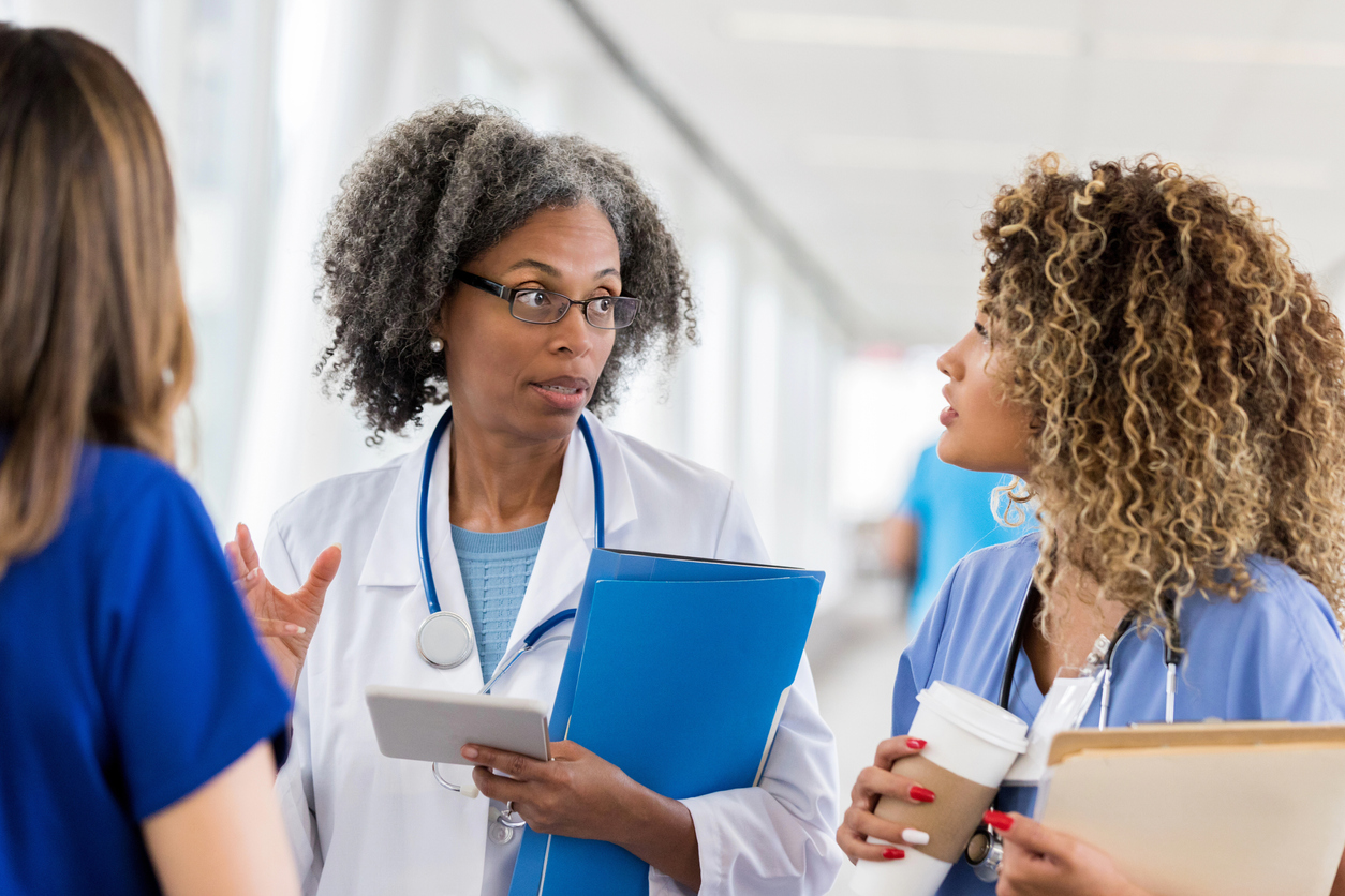 A nurse practitioner wearing a white coat talks with two nurses in scrubs.